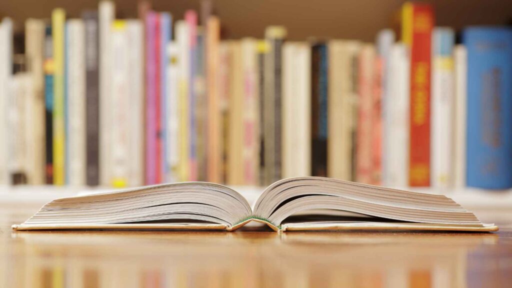 Im Vordergrund des Fotos liegt ein Buch aufgeschlagen auf dem Tisch. Weiter hinten steht ein Bücherregal voller Bücher.