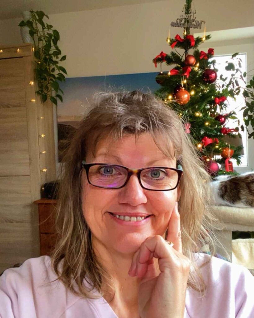 Ich lächle in die Kamera. Hinter mir mein diesjähriger festlicher Weihnachtsbaum. Auf dem Katzenbaum rechts im Bild schläft meine Katze.