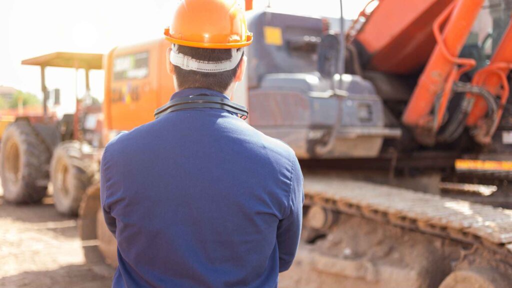 Ein Bauarbeiter beobachtet mit verschränkten Armen das Geschehen auf der Baustelle. Das Bild ist anonym, man sieht nur die Rückseite des Bauarbeiters und im Hintergrund Baumaschinen.