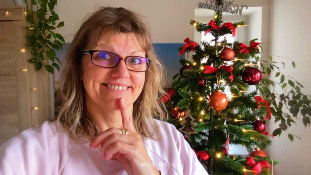 Ich selbst stehe im Wohnzimmer, hinter mir eine Lichterkette am Schrank und unser festlich geschmückter Weihnachtsbaum